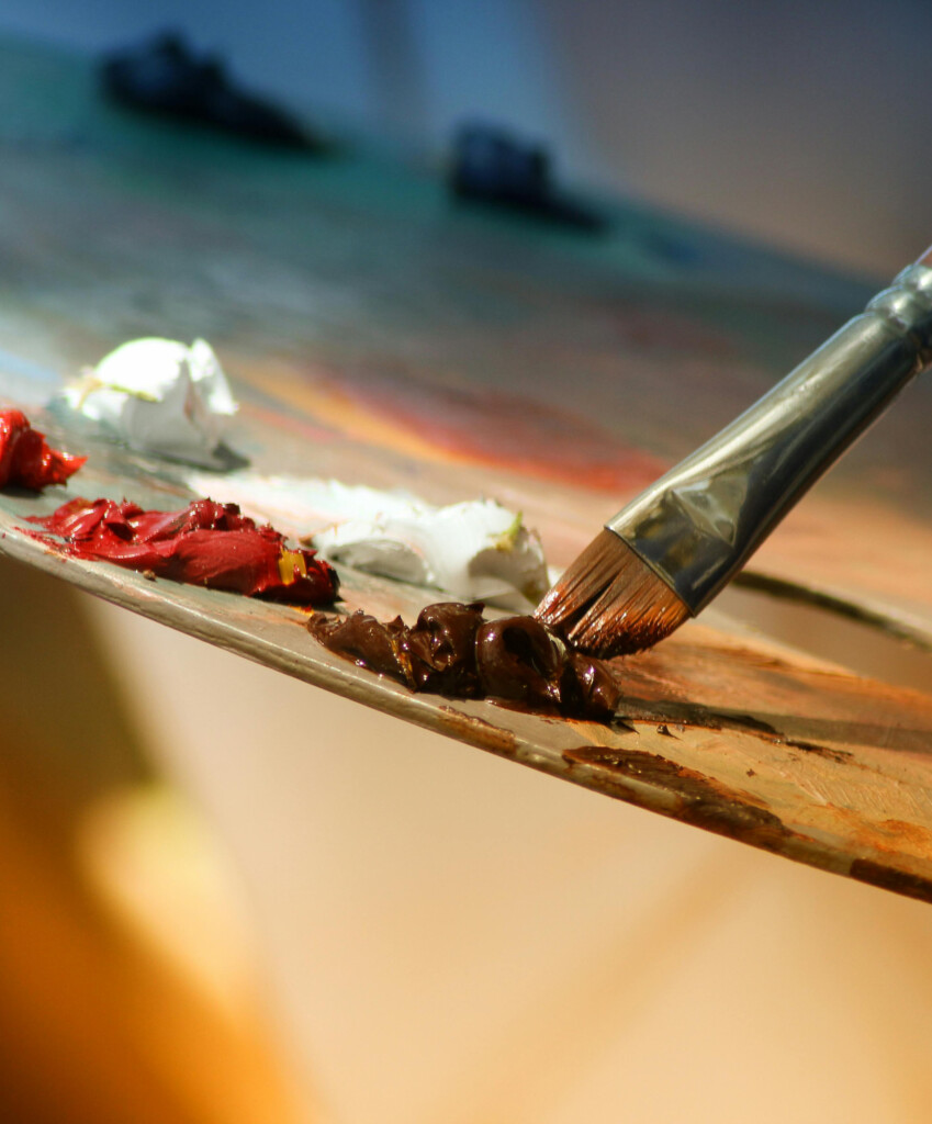 Close-up of a painter's palette with red, white, and brown paint blobs and a brush resting on the edge; blurred studio background.